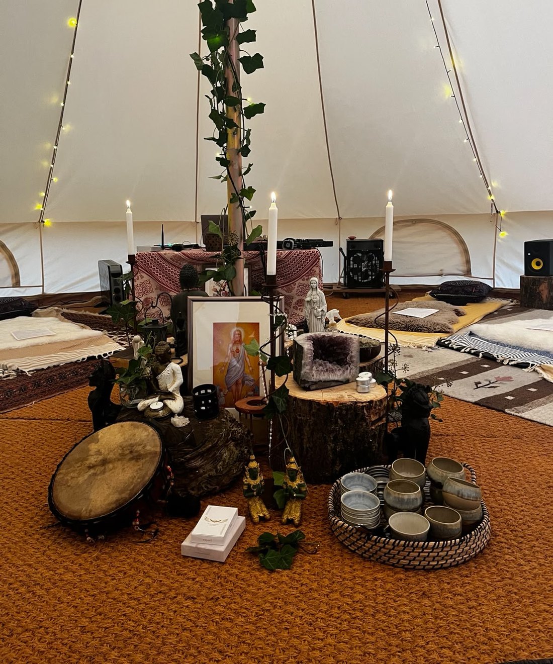 The altar inside the ceremony tent, photographed before a gathering: lit candles, a framed picture, small statues, an amethyst geode on a tree stump, a frame drum and a basket of ceramic cups, on a woven rug with sheepskins and cushions laid out in a circle around it.